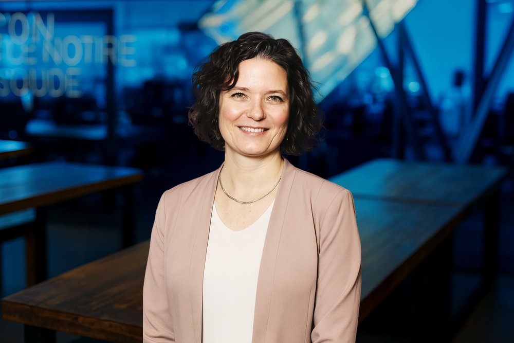 Sarah, directrice du déploiement agentique chez Vooban, souriante, veston rose poudré sur haut blanc, devant les bureaux de Vooban à l'éclairage bleuté