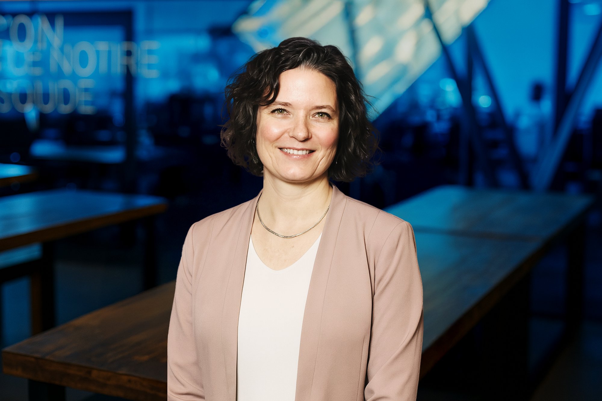 Sarah, directrice du déploiement agentique chez Vooban, souriante, veston rose poudré sur haut blanc, devant les bureaux de Vooban à l'éclairage bleuté